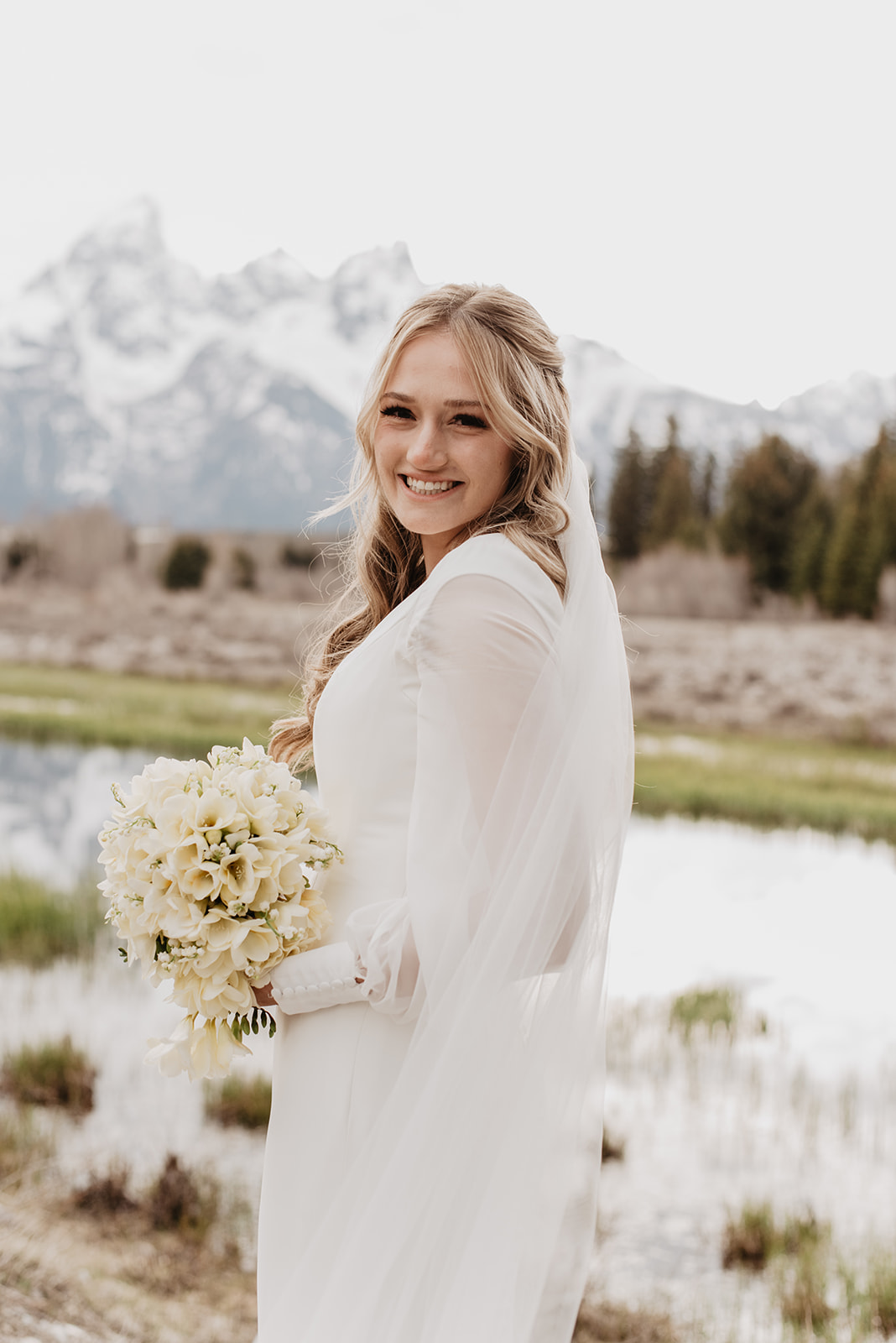 tall bride in a simple satin wedding dress looking over her shoulder and smiling at the camera with the Tetons behind her on her Jackson Hole wedding day