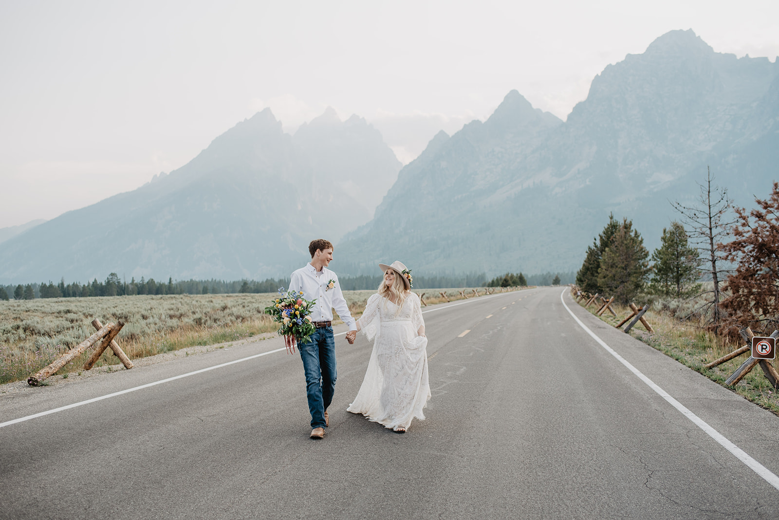 bride and groom walking across a road in Jackson HOle with the Tetons behind them as they hold has and smile at each other, 2022 wedding dresses