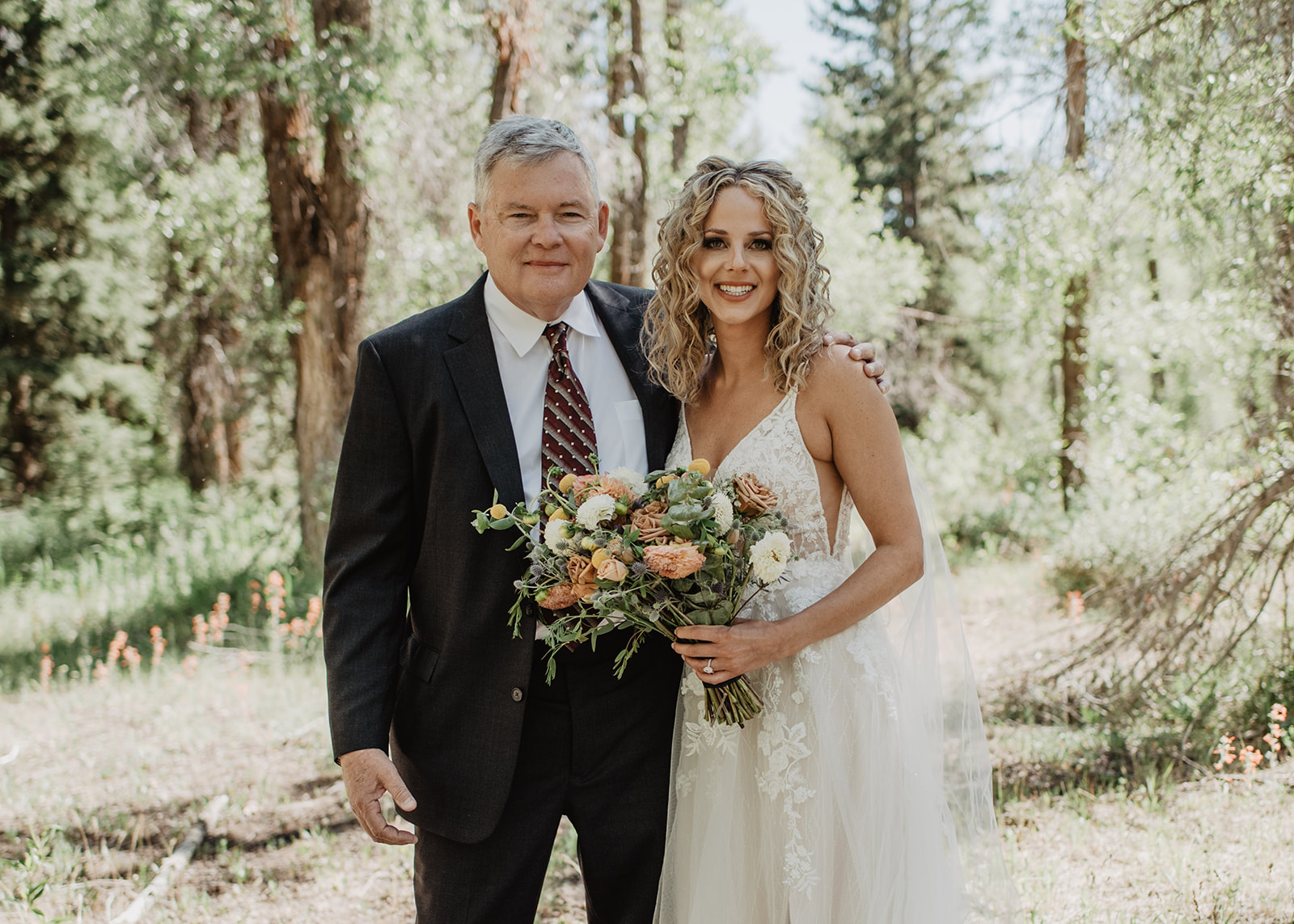 blonde bride in a lace dress standing with her dad in the woods in the Grand Tetons as she holds her bridal bouquet