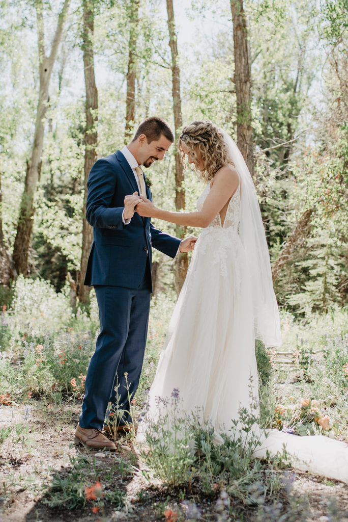 bride and groom holding hands together in JAckson HOle's forest as the groom looks at his brides lace white wedding dress