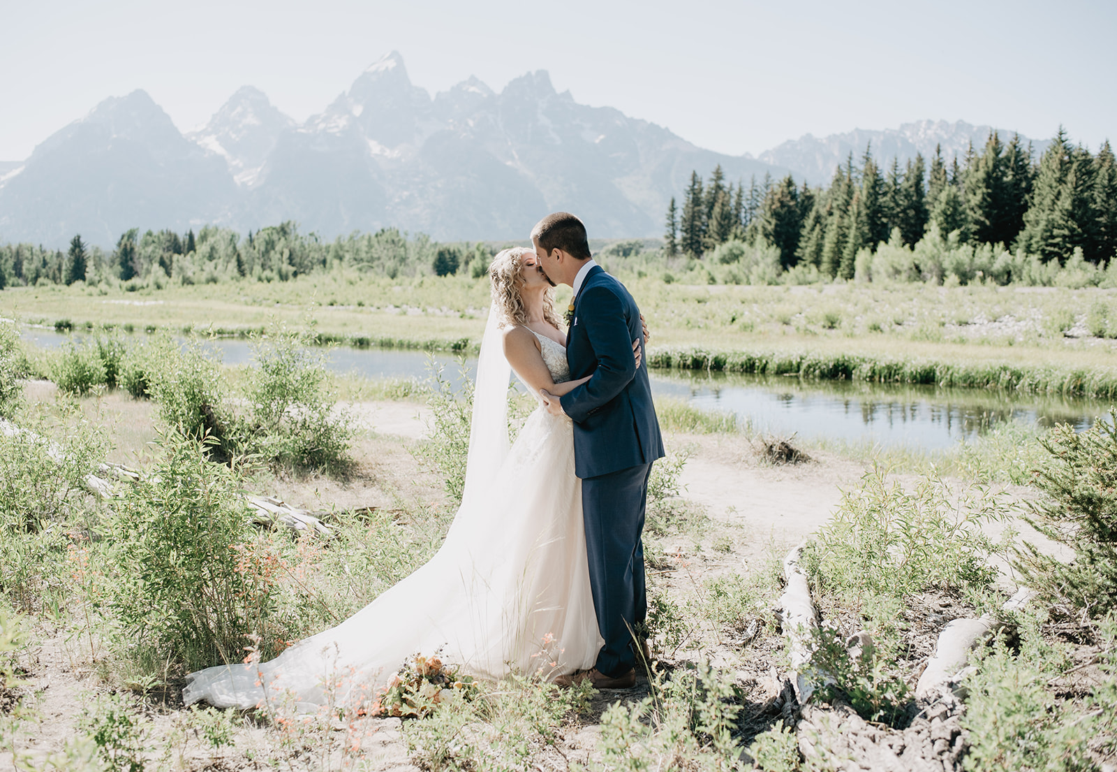 bride in bright white lace and tulle gown kissing her groom who is in a blue suit while they stand next to a river in Jackson Hole