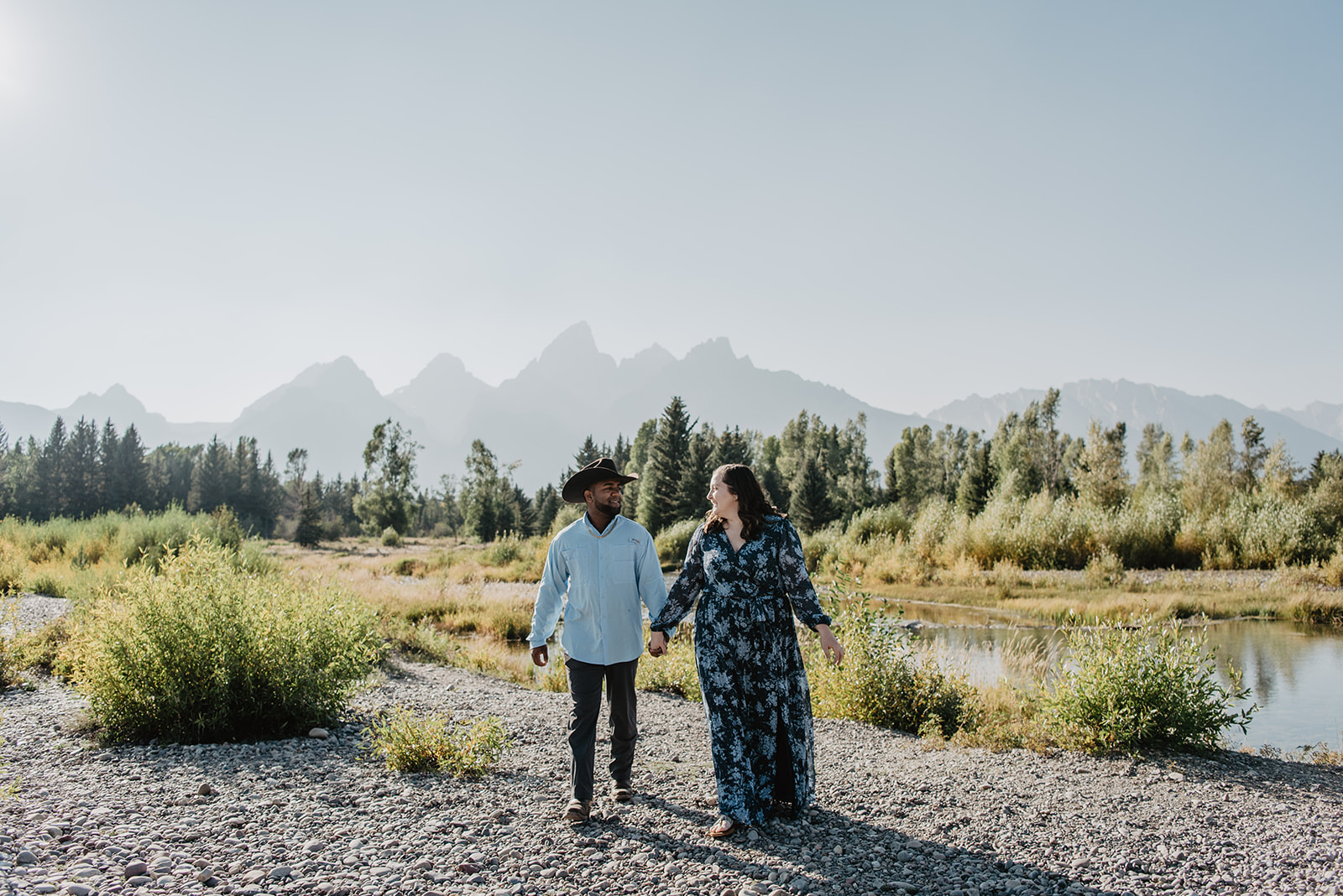 NAtional Park engagement photos with couple holding hands and walking a rock path towards the camera with the Grand Tetons mountain range behind them