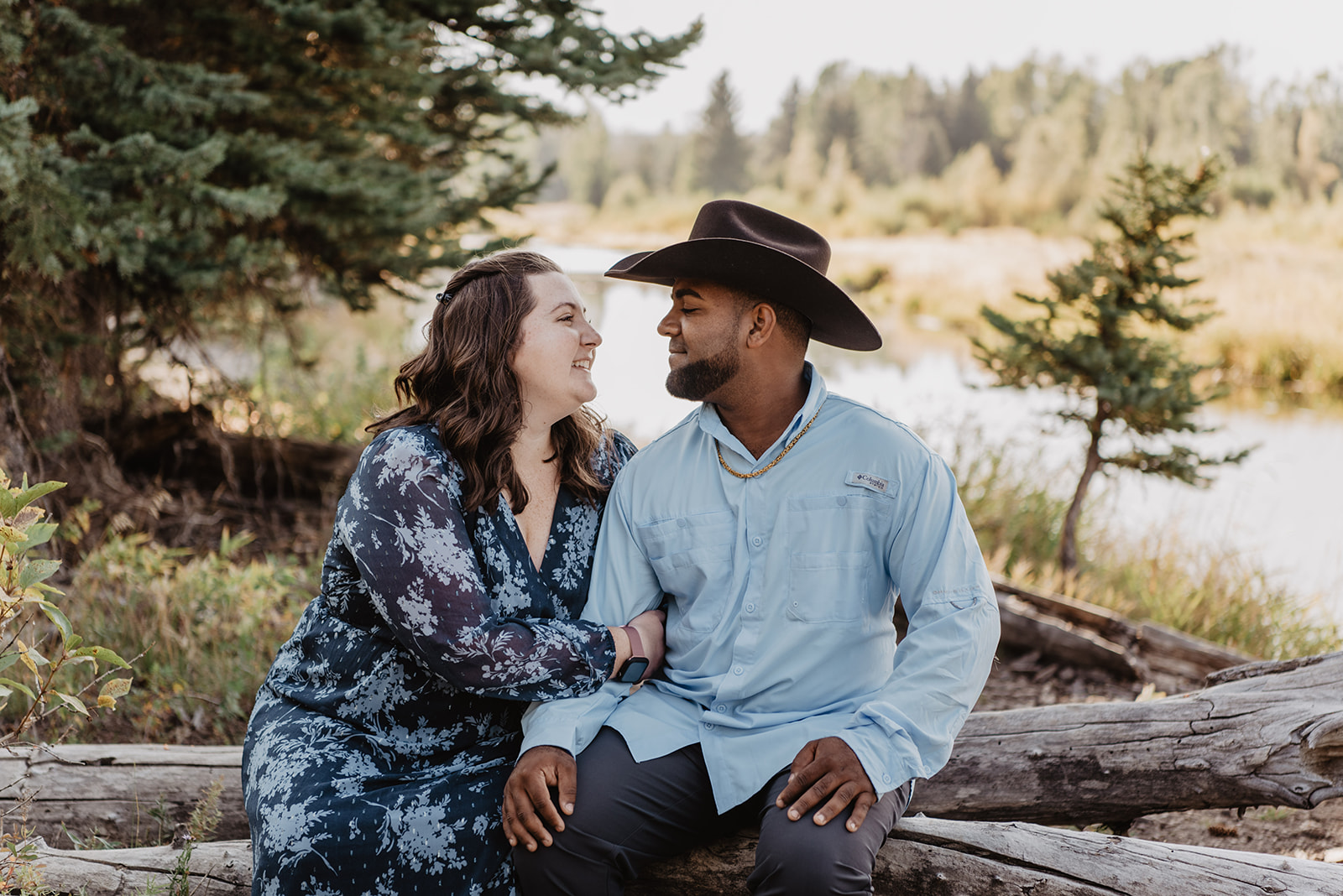 Jackson Hole engagement photos with man and woman sitting on a fallen tree with the water behind them looking into each others eyes romantically and smiling