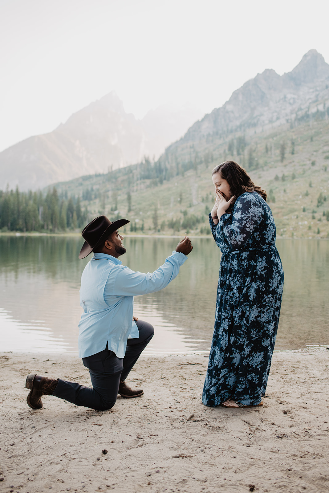surprise proposal at Jackson Hole with man in a blue button down shirt is on one knee proposing with a lake and the mountains behind them 