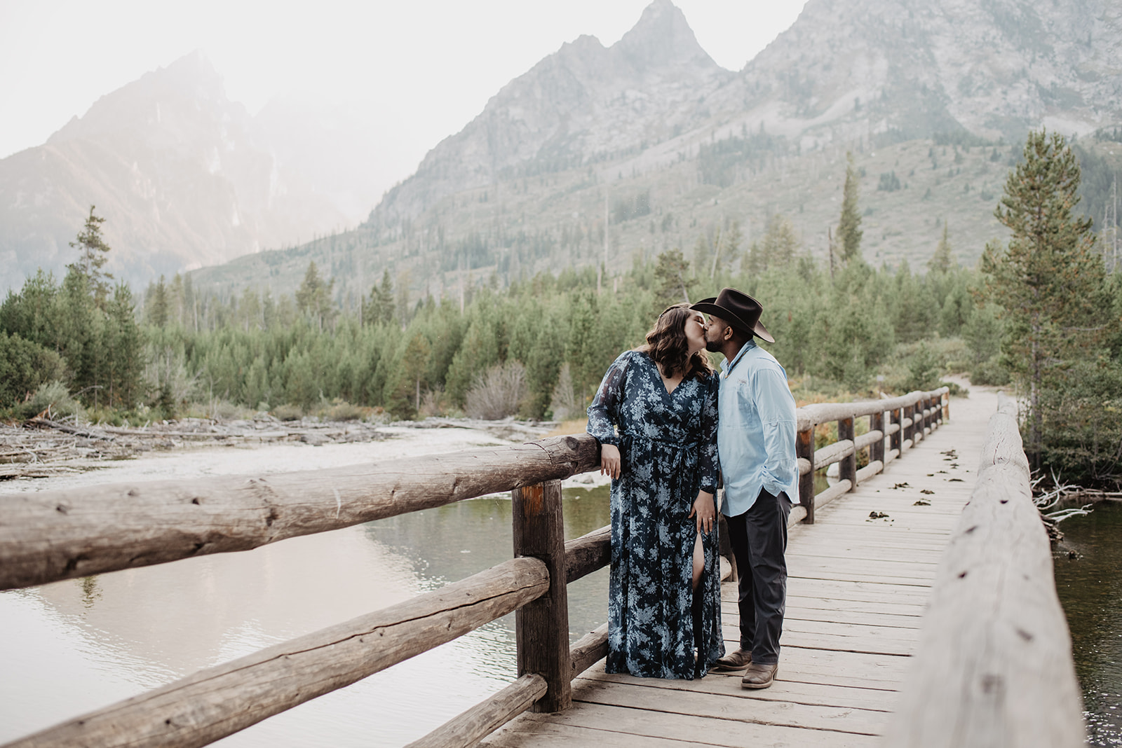 romantic western engagement photo outfits with man in a button down and cowboy hat and woman in a blue gown standing on a wood bridge in Jackson Hole kissing one another with the Grand Tetons behind them