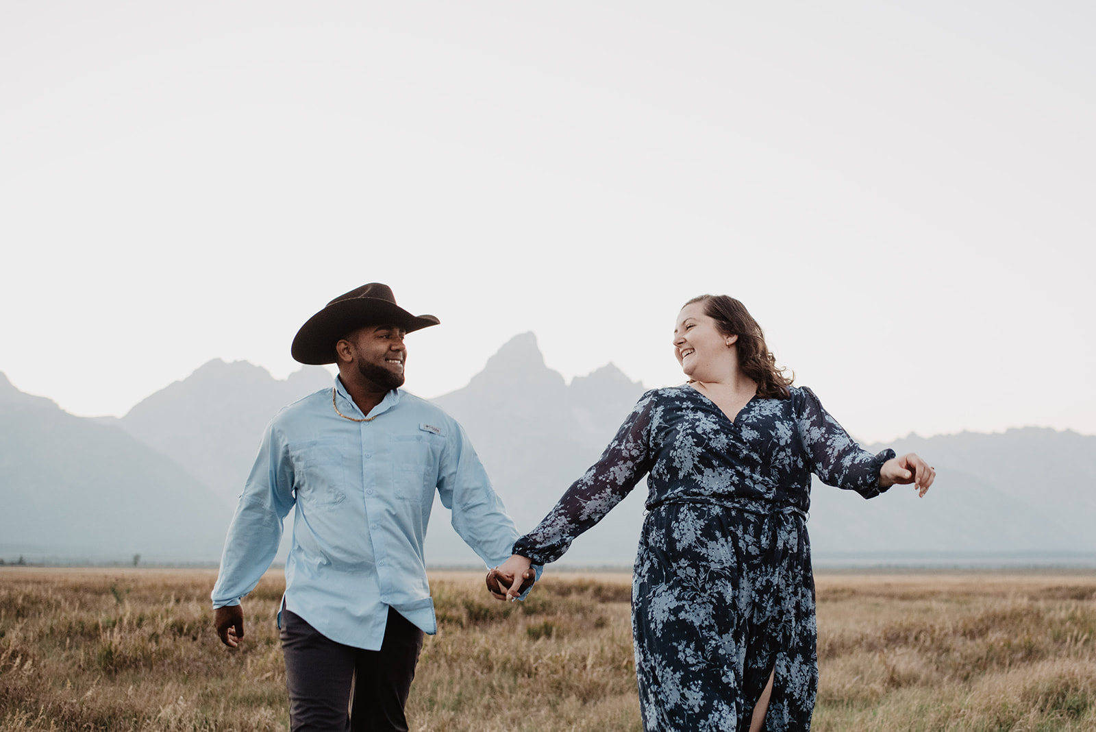 engagement photos in Jackson Hole with man and woman holding hands and running through a golden field with the Grand Tetons in the distance for an elegant western engagement photoshoot