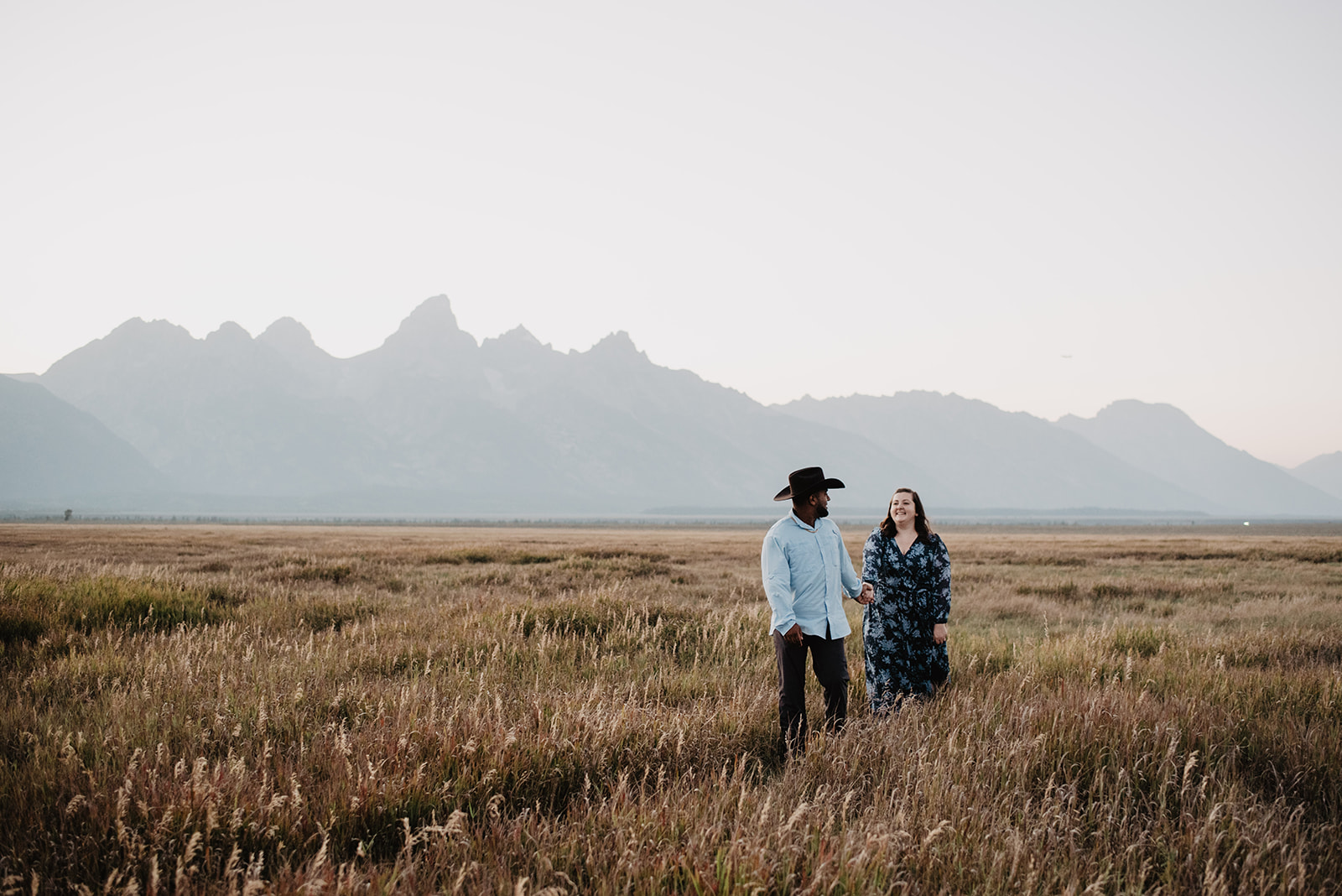 moody engagement photos in Jackson Hol with man and woman holding hands as they walk together through a meadow in the Grand Tetons on a hazy day