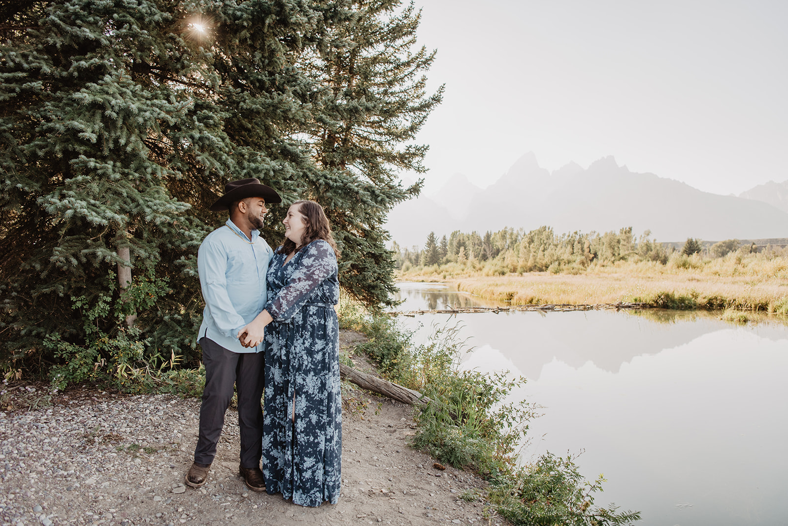 National Park proposal in the Tetons with a woman in a blue dress holding hands with her fiance next to a stream and the Grand Tetons behind them