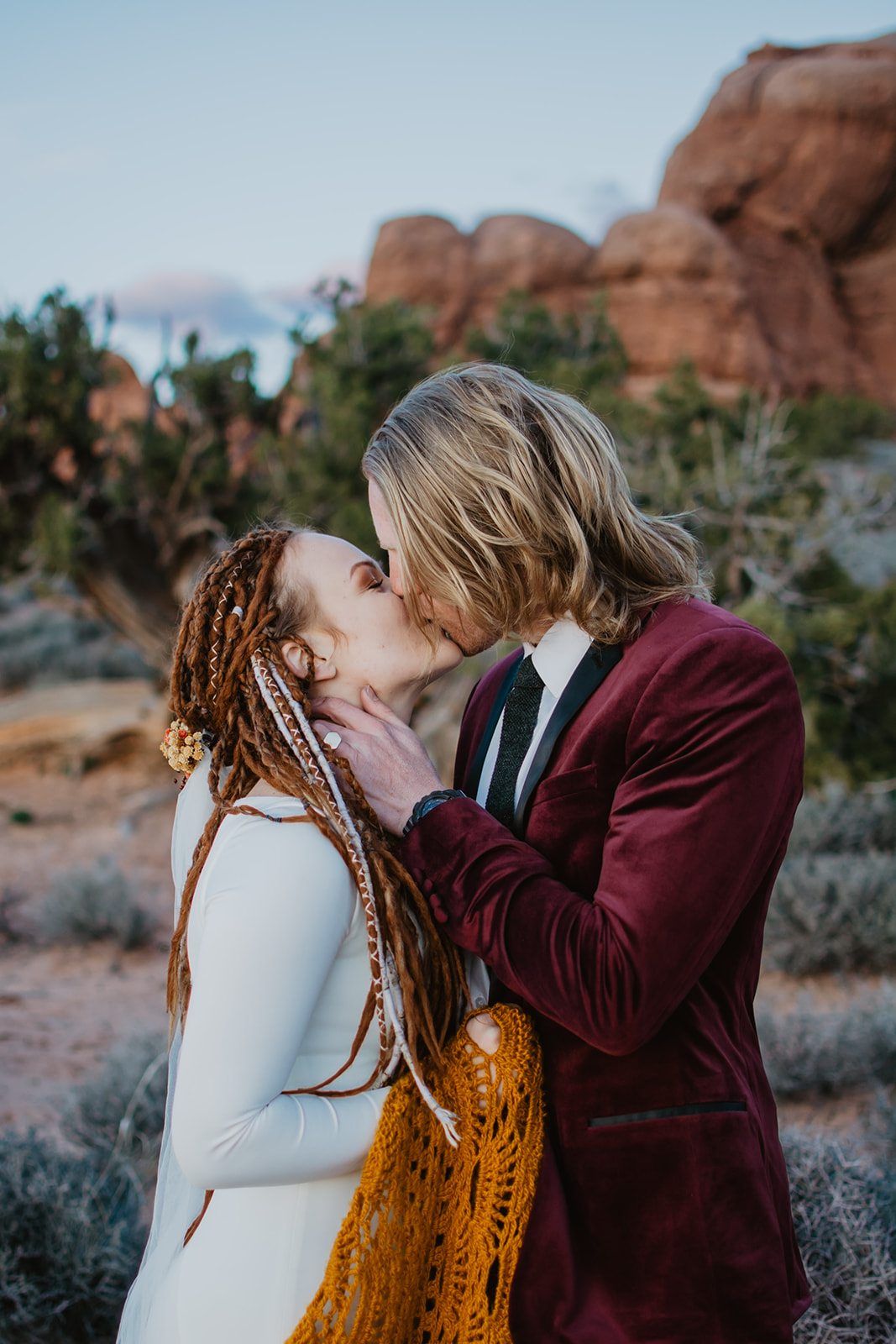 eloping in Moab with bride and groom passionately kissing each other with blue skies and the red rocks behind them, groom in a burgundy suit and bride covered in a burnt orange crochet shawl