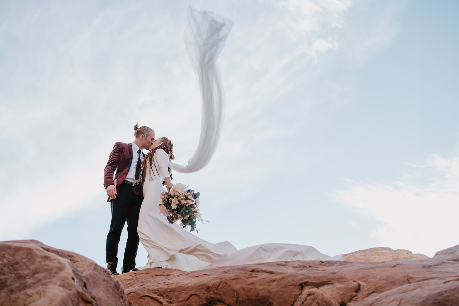 adventure elopement in Moab Utah with bride and groom hiking up to the top of an arch for an epic wedding photo with the wind blowing the brides veil while the couple kisses