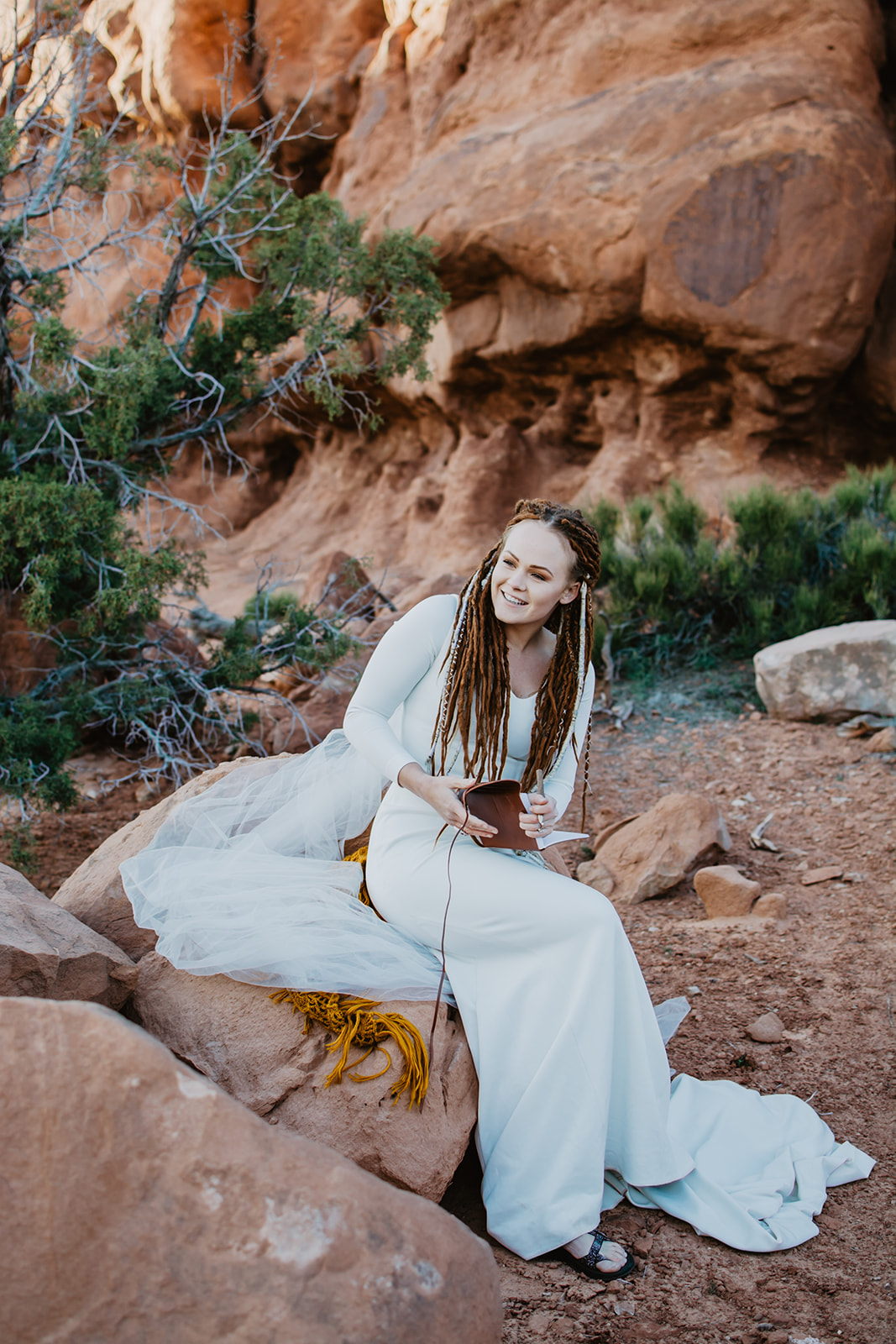 beautiful bride with red haired dreading and a long sleeved satin wedding gown sitting on a rock in the red rock desert writing her vows