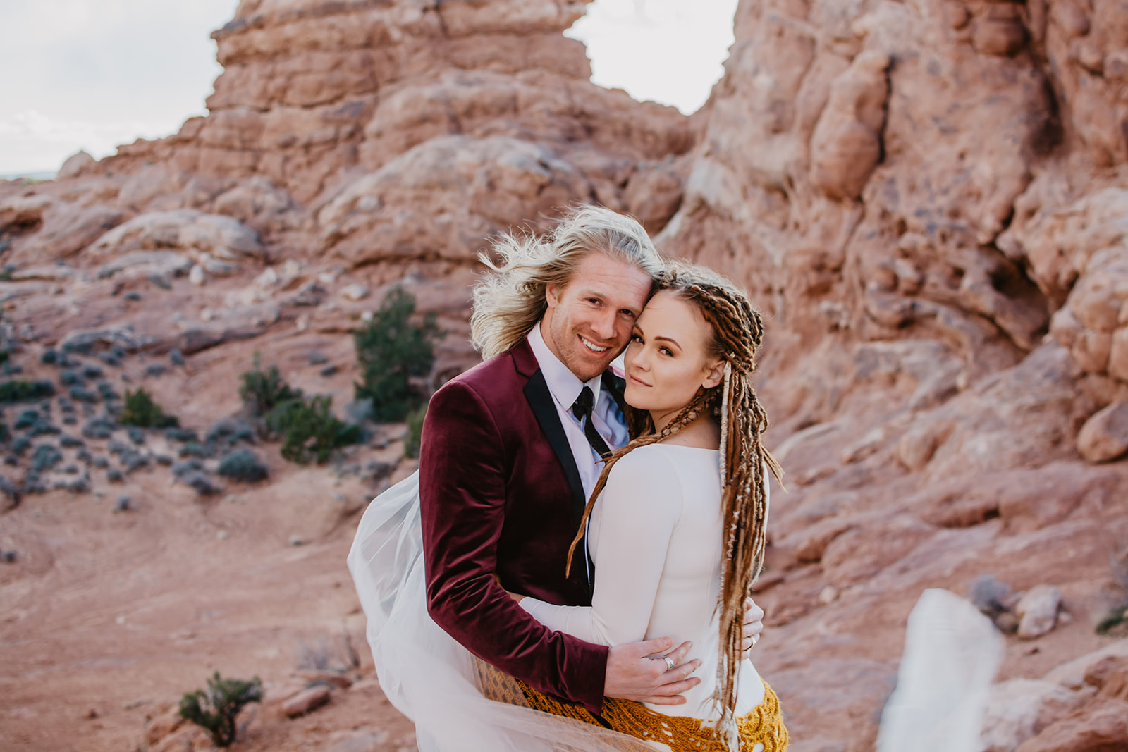 red rock elopement with bride and groom hugging and smiling at the camera with the rocks and shrubs in the background captured by the best Moab elopement photographer