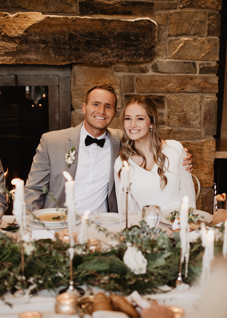 bride anda groom sitting down for dinner at the Four Seasons in Jackson Hole with a gorgeous fireplace behind them and greenery and candles decorating the table