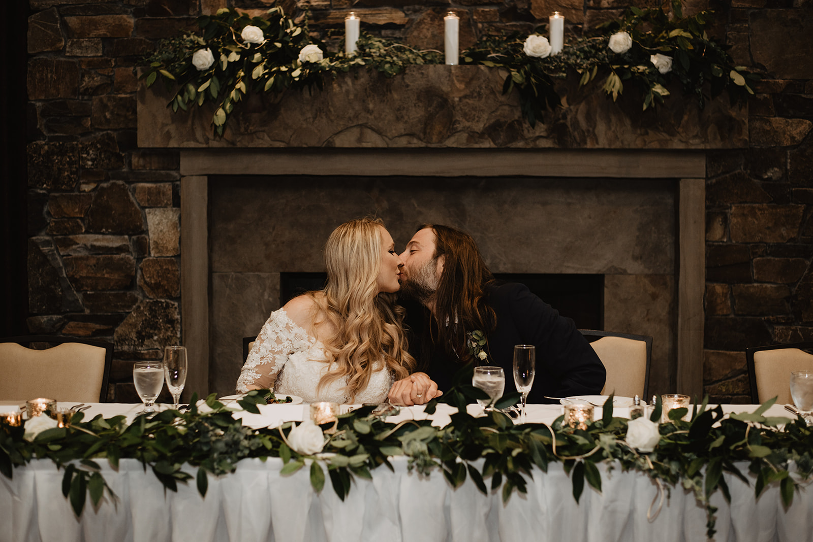 bride and groom kissing as they sit at their table that is covered in a white table cloth and magnolia leaves