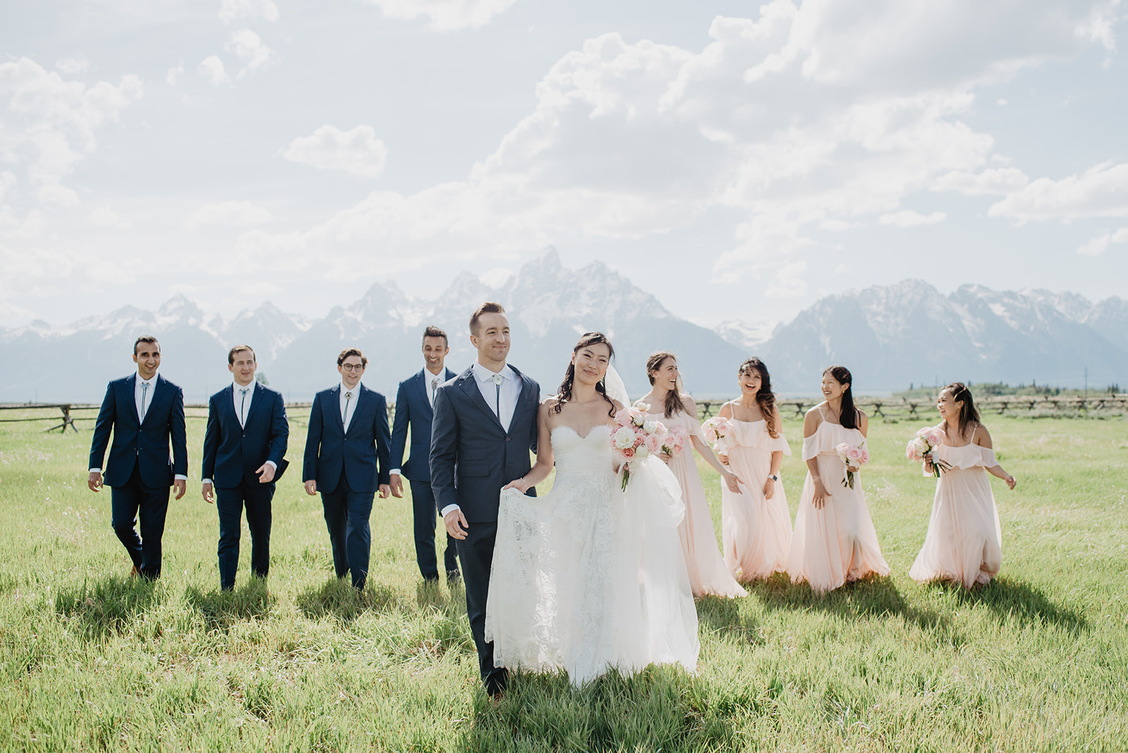 bride and groom holding hands and walking to the camera with their bridal party behind them in a green field