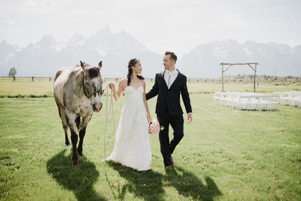 bride and groom walking together in a green field while leading a horse with the Grand Tetons in the background