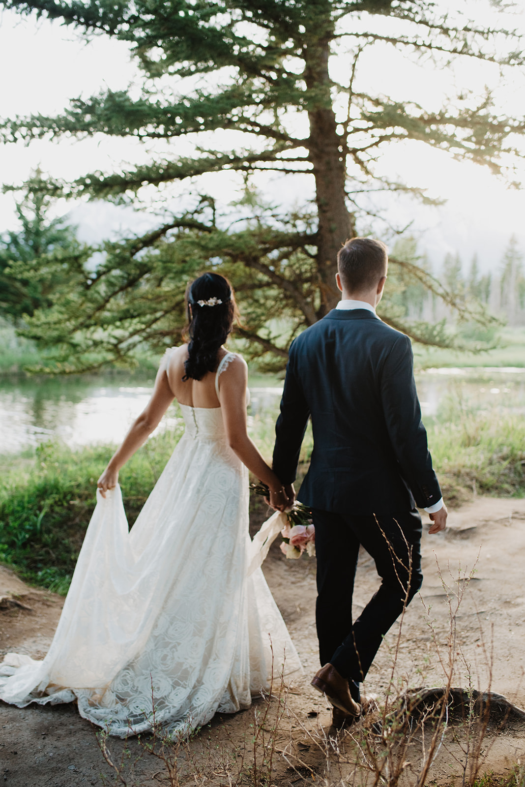 bride and groom walking on a dirt path in Jackson Hole next to a river as they hold hands and walk away from the camera, captured by the best Jackson Hole wedding photographer
