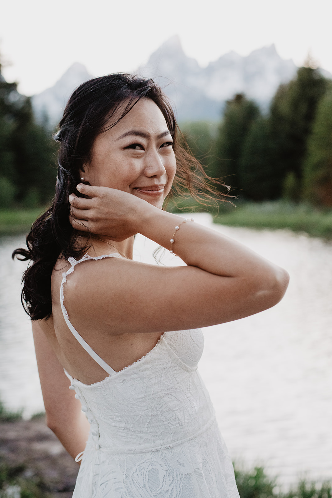 beautiful bride standing next to a river in the Tetons as she looks over her shoulder at the camera and smiles with the Tetons behind her