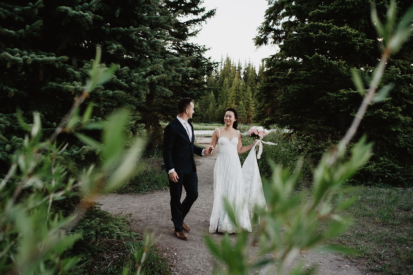 adventurous bride and groom have a wedding photo session in the Grand Tetons for their destination wedding day as they hold hands and walk down a trail together at sunset in the mountains