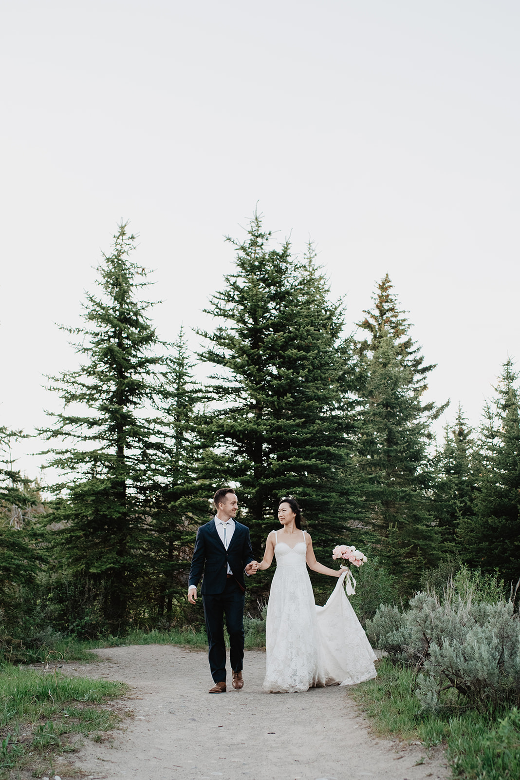 extended wedding photo shoot with Jackson Hole bride and groom holding hands and looking at one another as they stroll down a path with pine trees behind