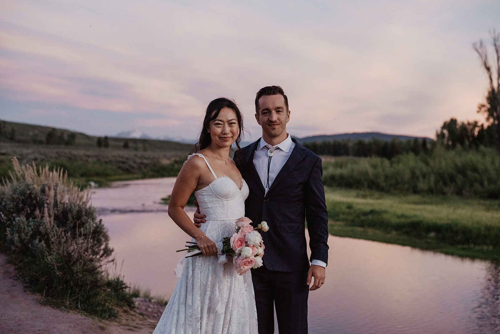 sunset wedding photos in the mountains of the bride and groom standing on the river with the pink sunset behind them