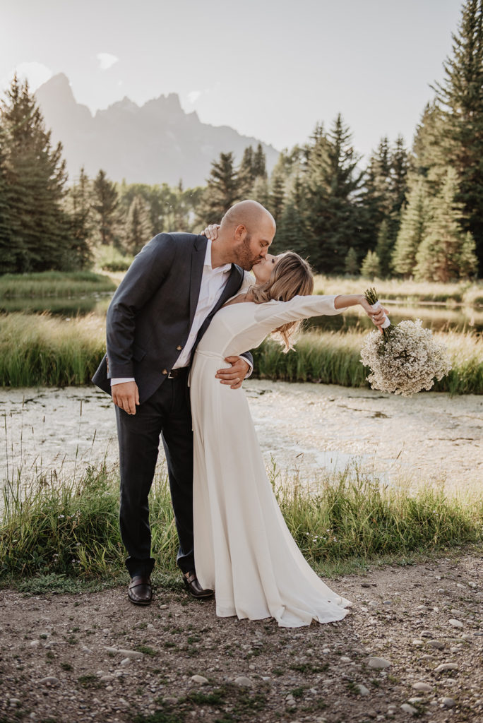 Jackson Hole photographers capture bride and groom kissing in front of a river at the end of their wedding ceremony in the grand tetons 