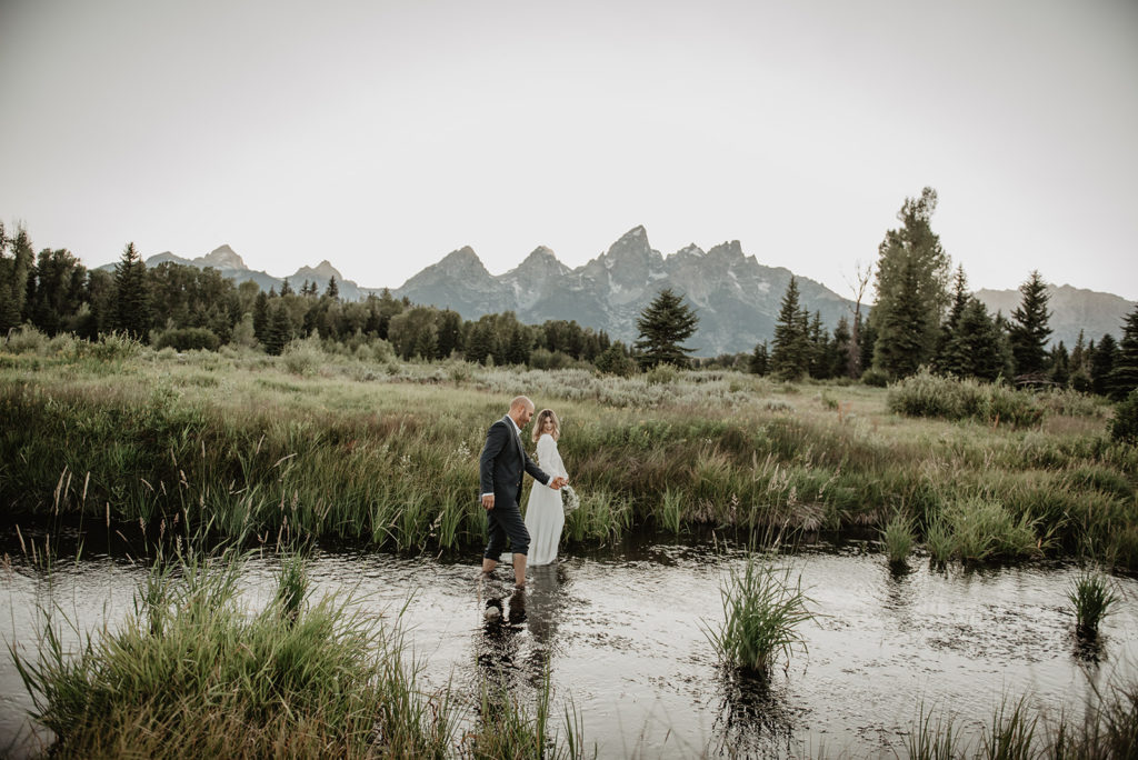grand teton wedding photographer captures bride and groom holding hands and walking through a river at Schwabacher landing with the tetons in the distance 