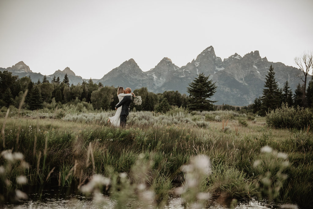 Jackson Hole photographer captures bride and groom in the Tetons with the groom picking up his bride and spinning her around in a sage brush field with the mountains in the distance