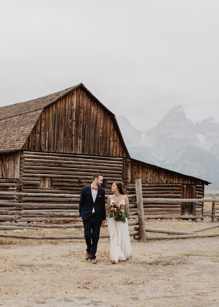 bride and groom holding hands and walking away from an old historic barn with the Tetons in the distance photographed by Jackson Hole wedding photographer 