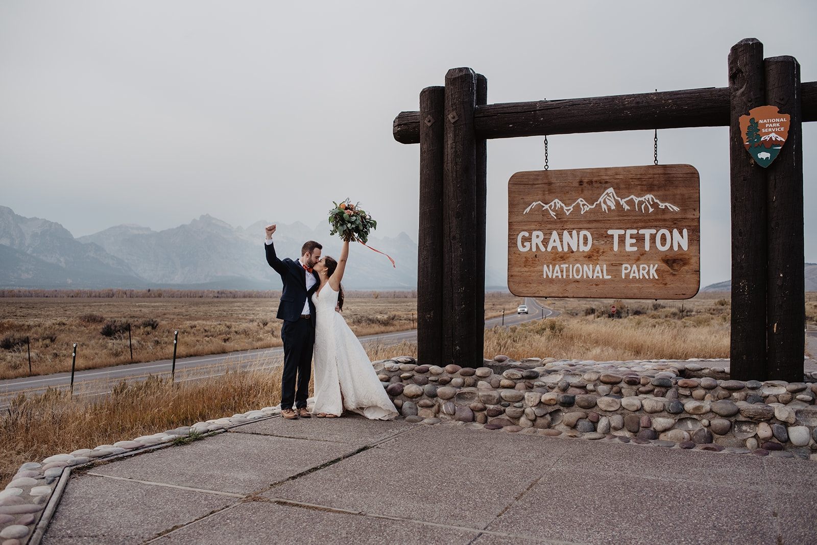Bride and groom standing next to the Grand Teton National Park sign with their arms in the arm in celebration