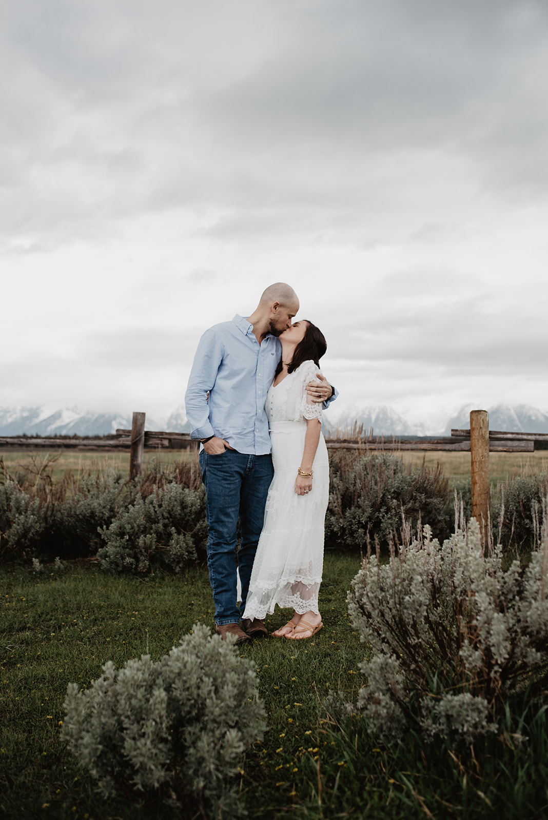 rainy day photos in the Tetons with husband and wife taking anniversary photos in a sage brush field with a wood fence behind them