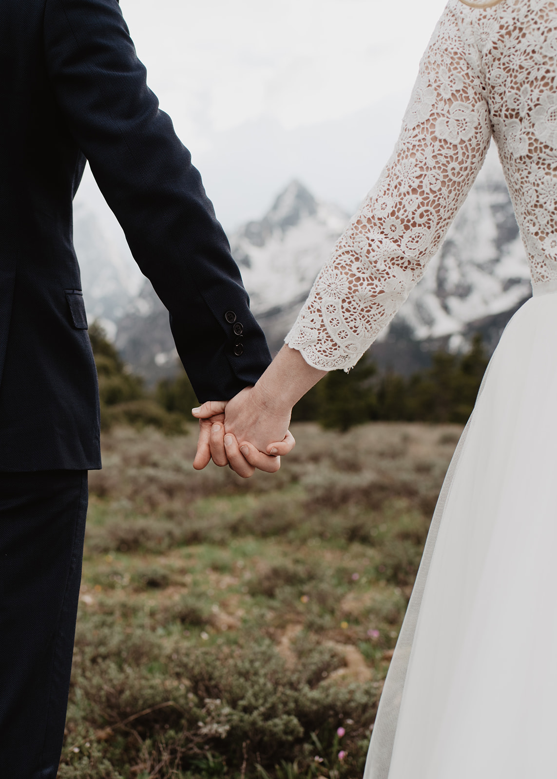 detail shot of bride and groom holding hands as they walk through a field for their Grand Teton Spring elopement captured by best Grand Teton Wedding Photographer