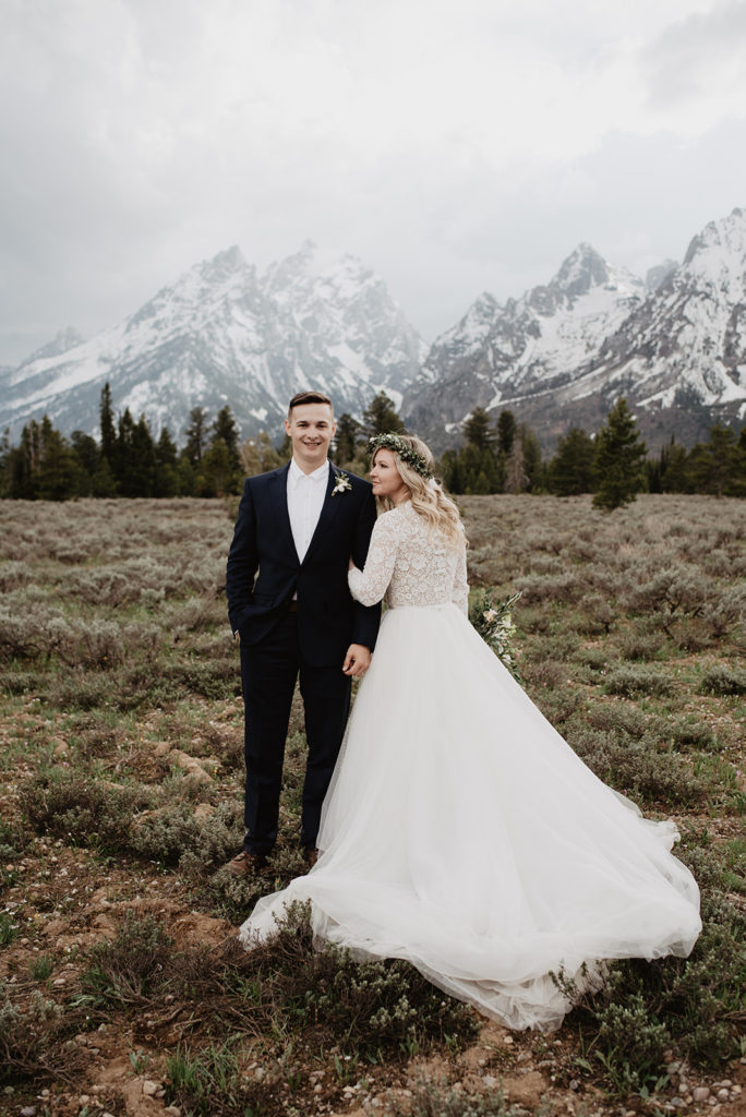 Jackson Hole wedding photographer captures bride and groom holding hands and standing together in a field in front of the mountains on a cloudy day
