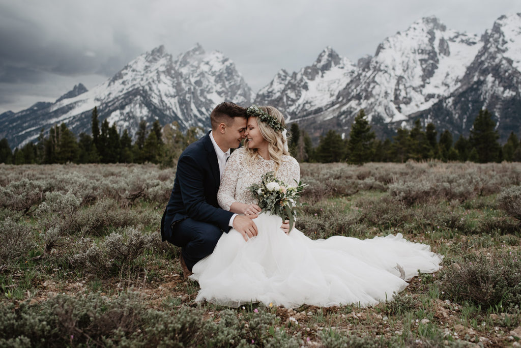 bride and groom sitting in a field together with the snowy Grand Tetons right behind them with dark clouds rolling into the field