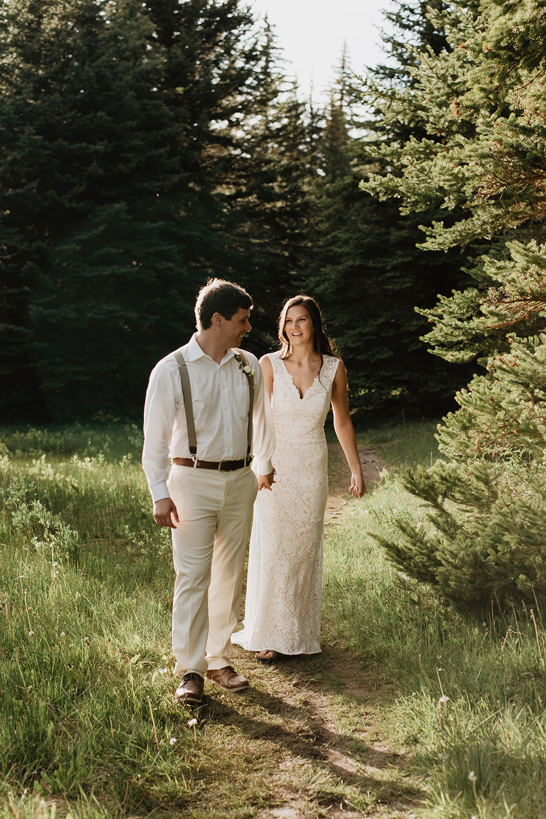 groom holding his brides hand and leading her down a dirt path past a forrest of trees in the Grand Tetons as he looks back at her and smiles 