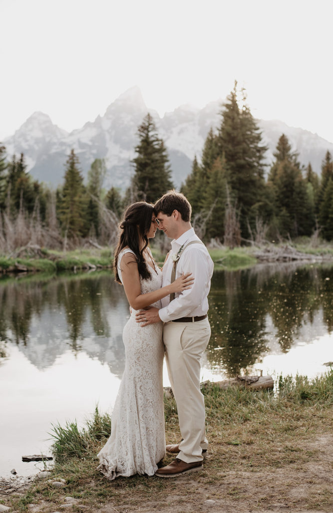 bridal portraits with bride and groom embracing one another while resting their foreheads together with the Grand Tetons in the distance reflecting over a small lake captured by Jackson Hole photographer 