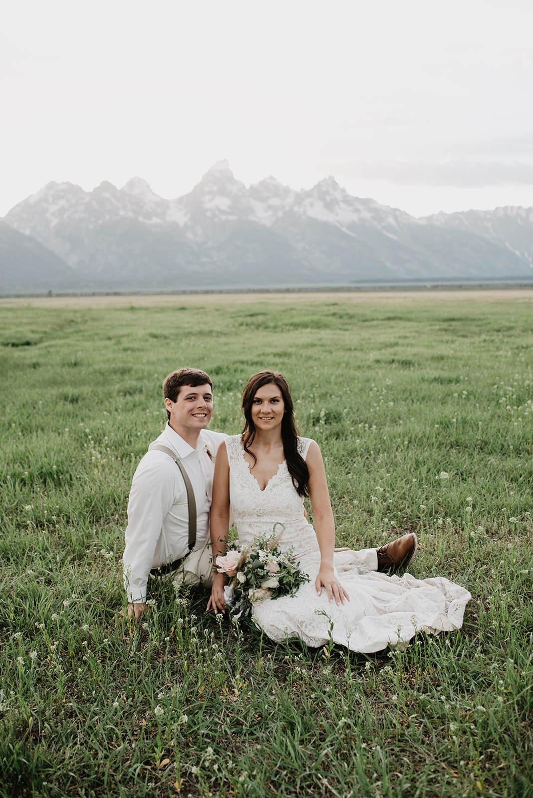 bride and groom sitting in a green field together with the Tetons behind them for their Jackson Hole summer wedding 