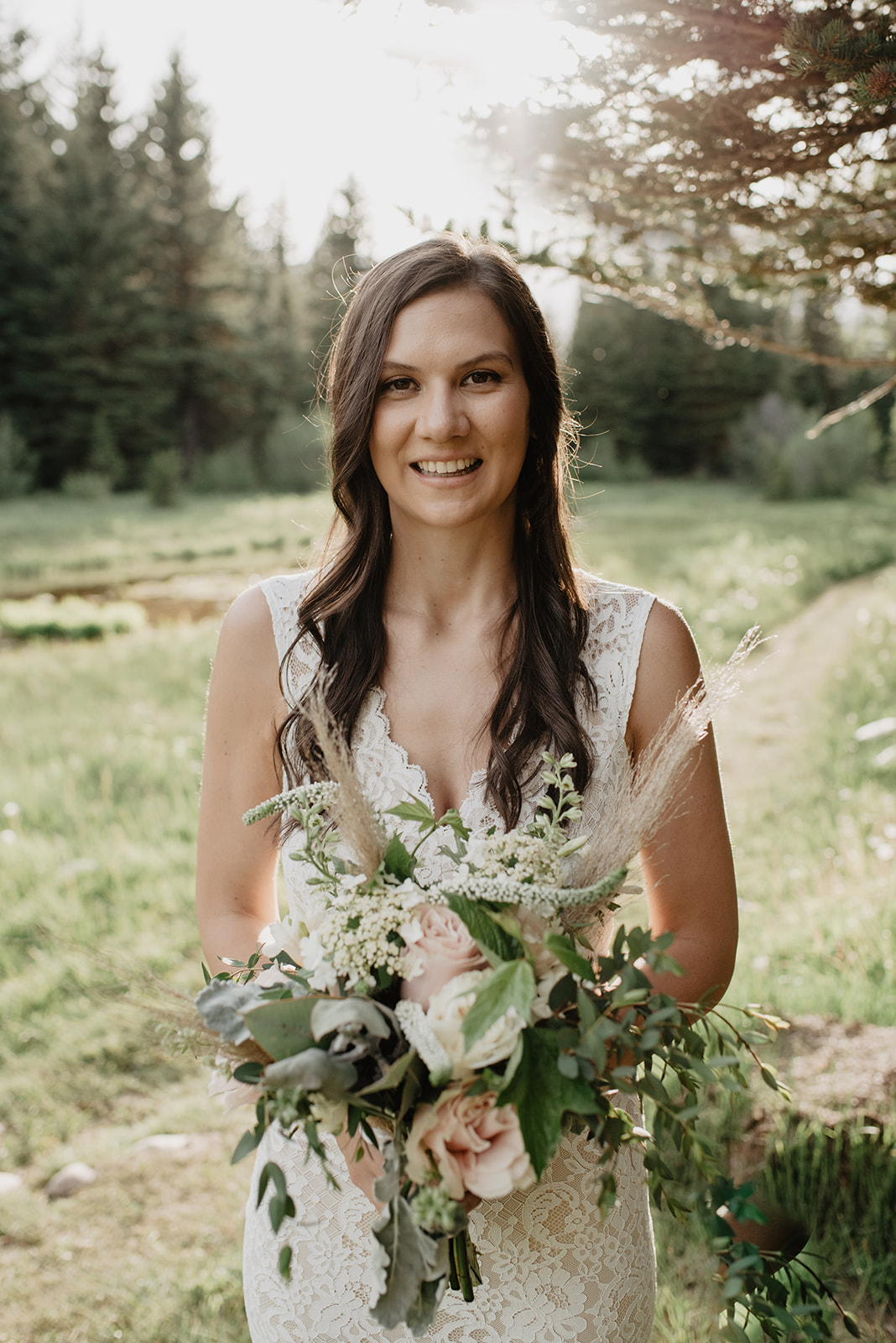 bride in a lace v neck wedding dress smiles as she holds her blush pink florals on her wedding day in Grand Teton National Park