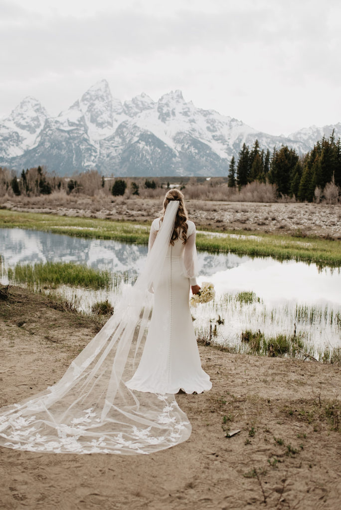 bride standing on the shore of a river in the Tetons with her cathedral veil trailing behind her on the ground as she looks at the snowy Tetons in the distance captured by Jackson Hole photographer