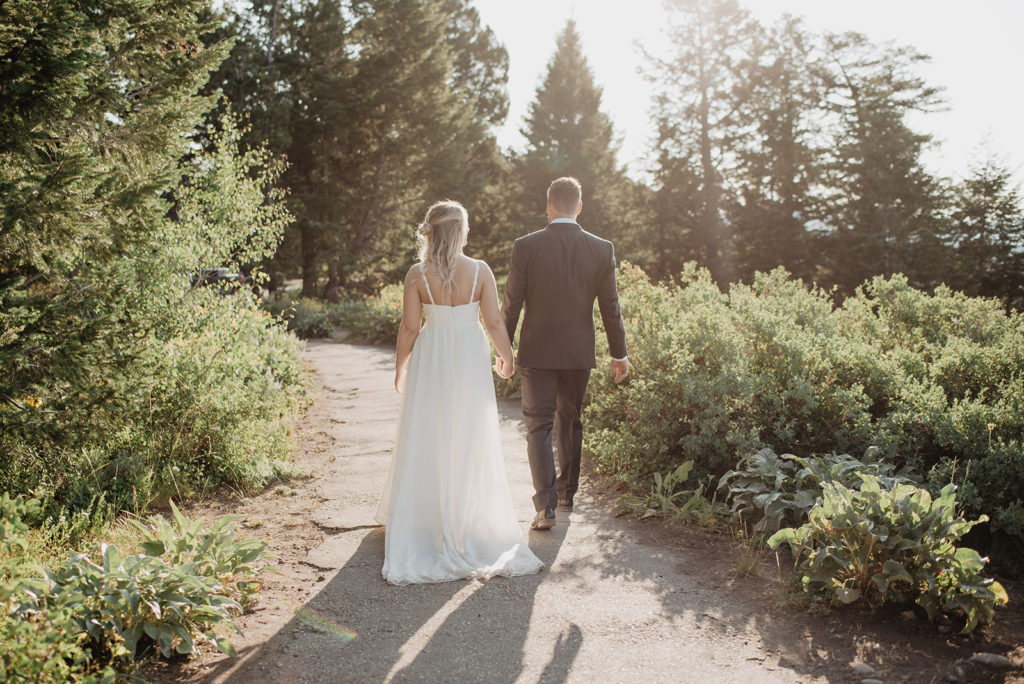 bride and groom holding hands and walking up a trail in the woods while the sunsets over the tree line photographed by jackson hole photographer