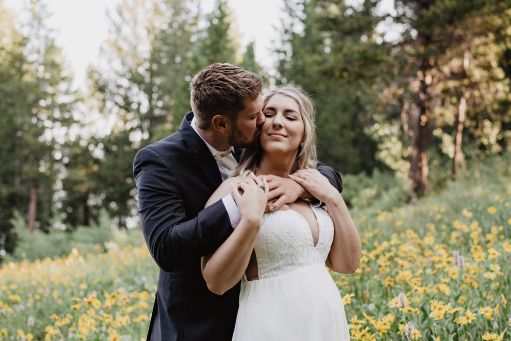 groom standing behind his bride and wrapping his arms around her shoulders as he learns in to kiss her on the cheek