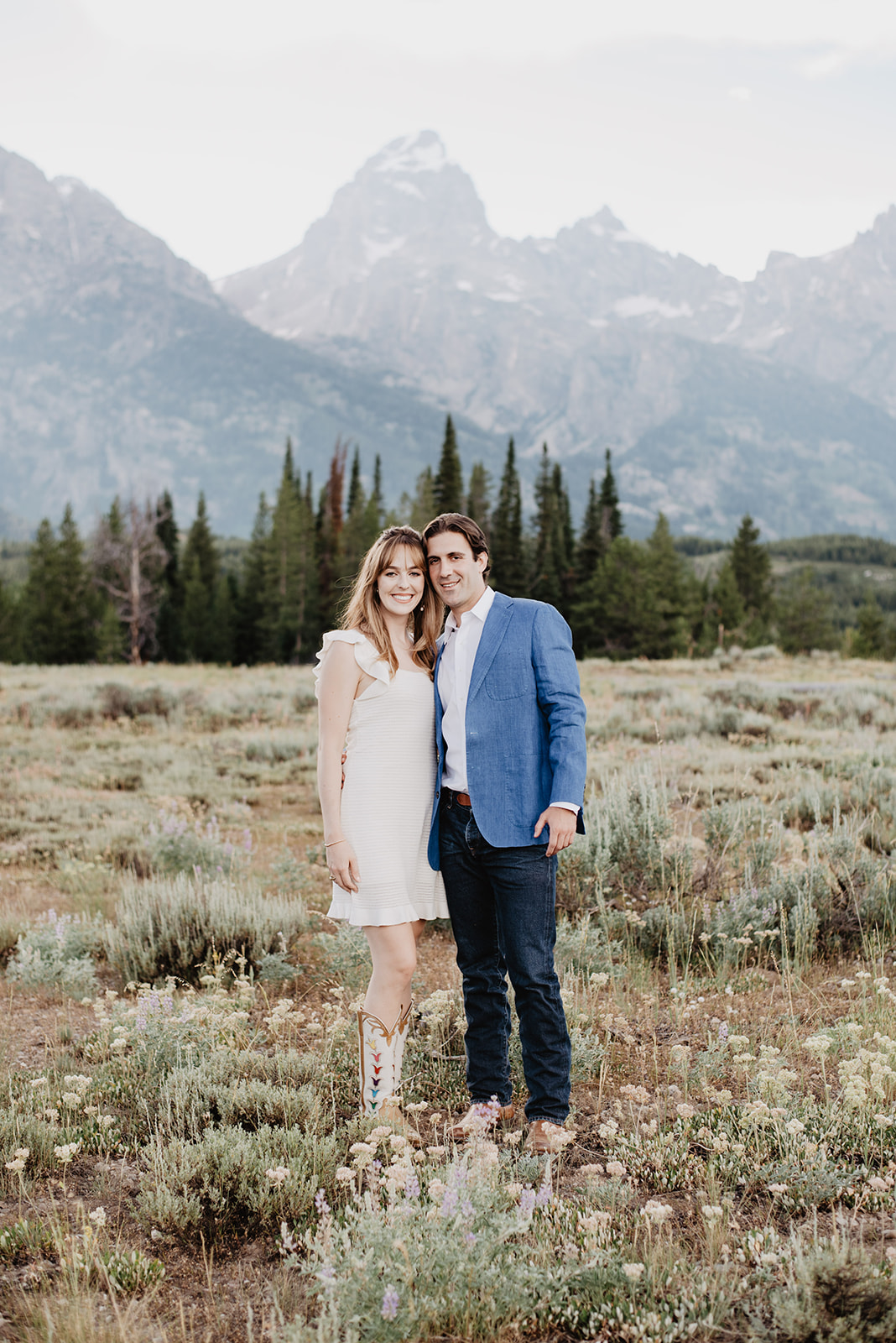 engagement portrait of man in a blue blazer and woman in a white dress standing together in Jackson Hole