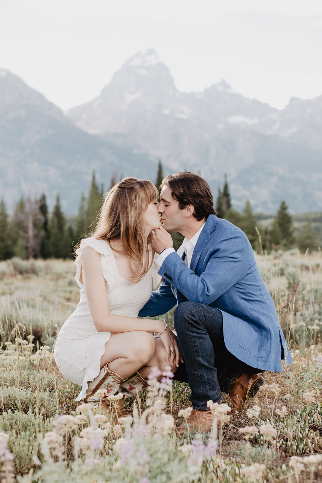 Jackson Hole photo tour engagement session with man and woman sitting in a meadow with the Tetons behind them as they kiss each other in their formal engagement photo outfits