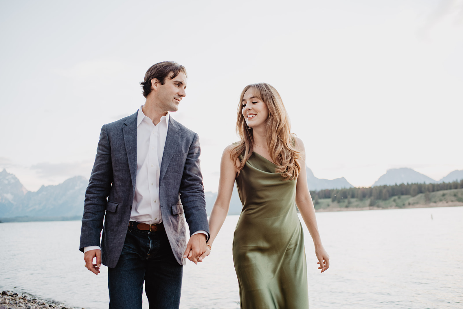 man in a grey blazer and womanin a green cocktail dress hold hands and walk along the breach on a lake in Jackson Hole photo tour engagement session