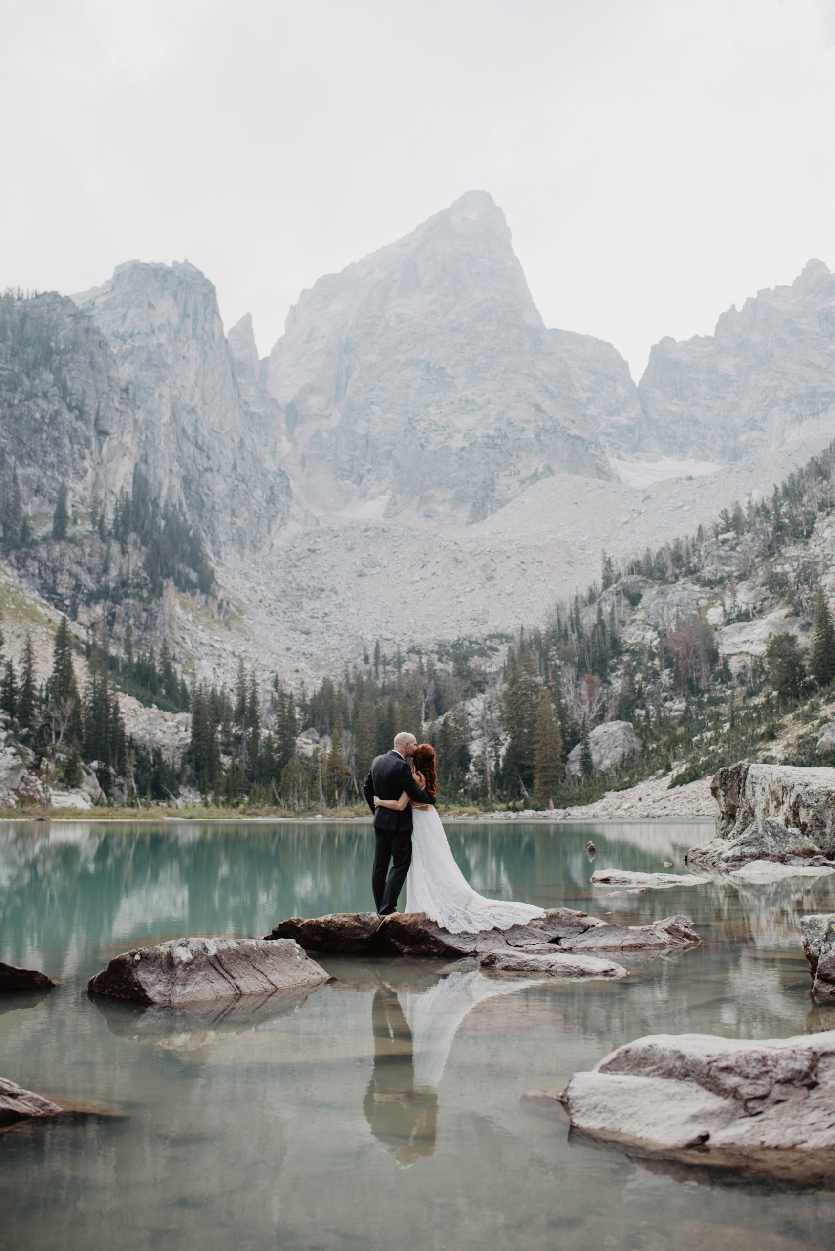 bride and groom elope in the mountains with Grand Tetons NAtional PArk wedding photographers at Glacier Lake, bride and groom standing on a rock in the middle of the lake with the mountains towering over them 