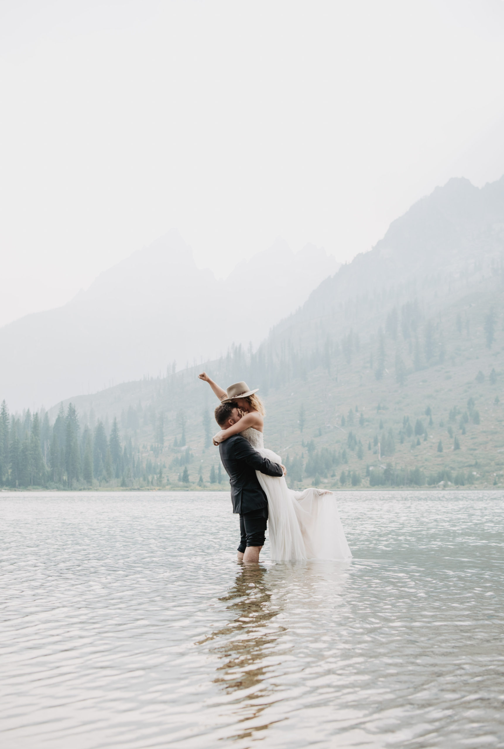 bride and groom standing in the water in a lake in Grand Tetons for their wedding with groom carrying bride as she punches the air in celebration