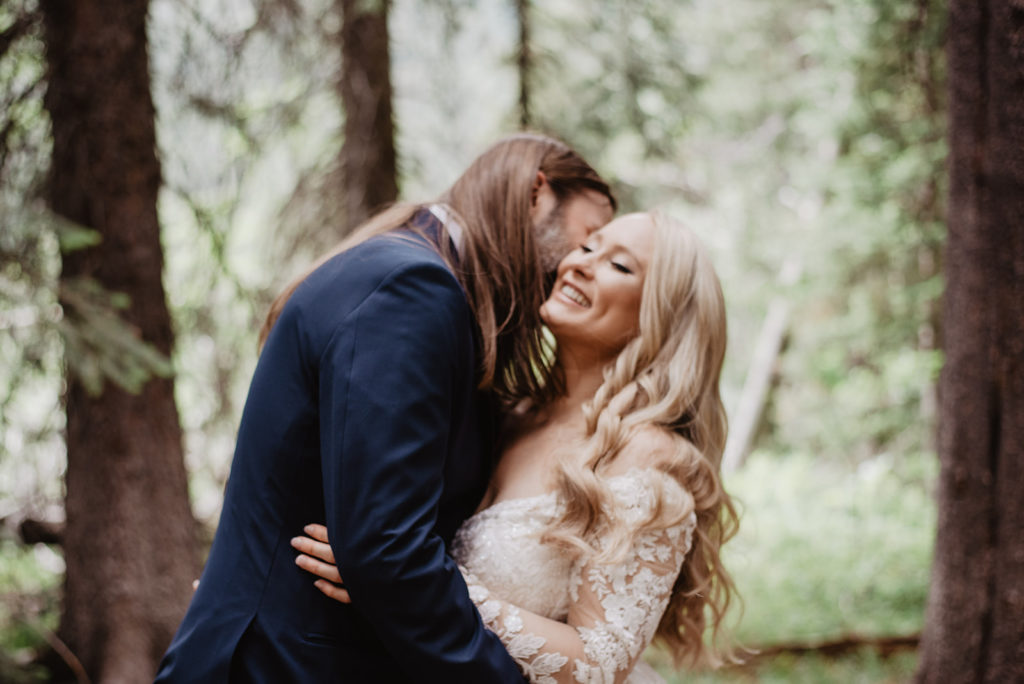 groom hugging his bride while he kisses her cheek as she looks into the sky and smiles while they are in the woods in Jackson Hole