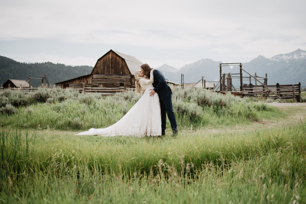 bride and groom kissing in a green field in Jackson Hole in front of an old barn at Mormon Row for their summer wedding 