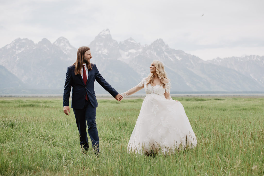 Jackson Hole destination wedding with bride and groom holding hands and running through a green field with the mountains in the background