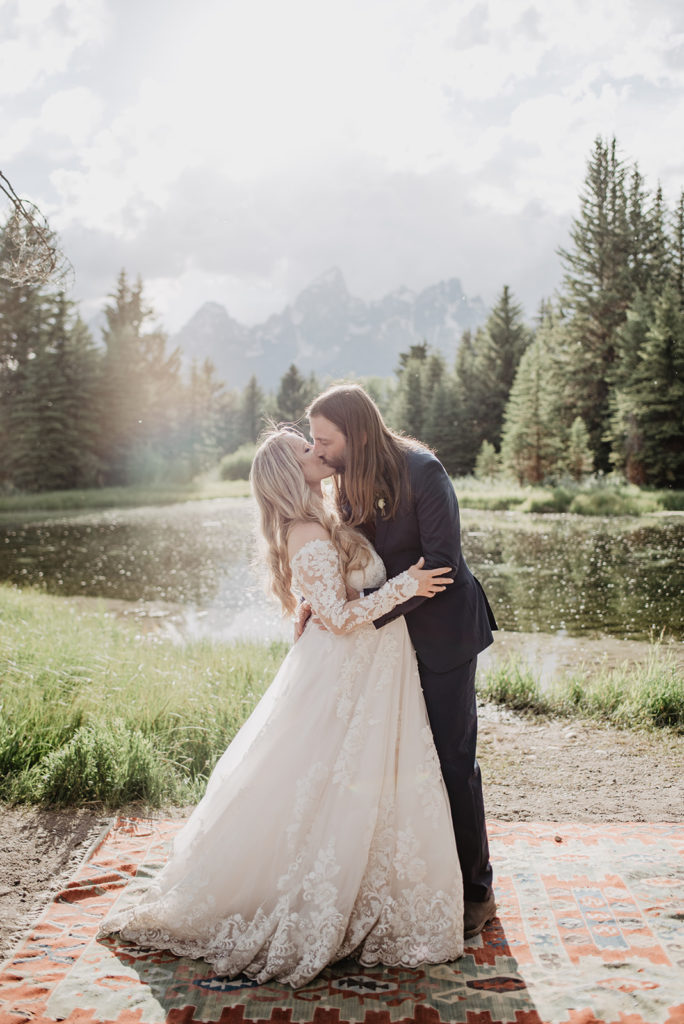 groom dips his pregnant bride back and kisses her passionately in front of a river with a forest and mountains in the distance 